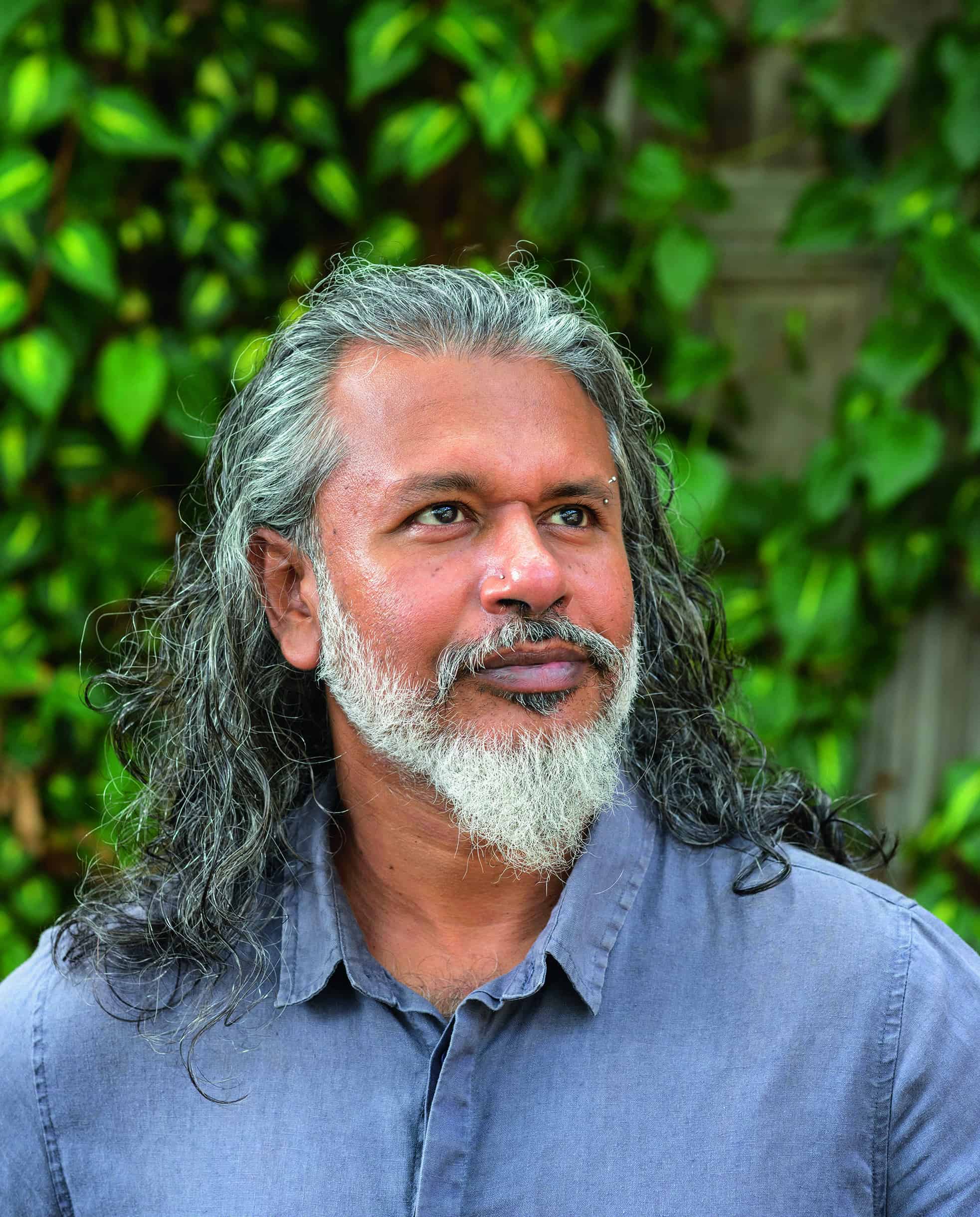 Shehan Karunatilak - a long-haired, bearded South Asian man in his 40s wearing a blue shirt