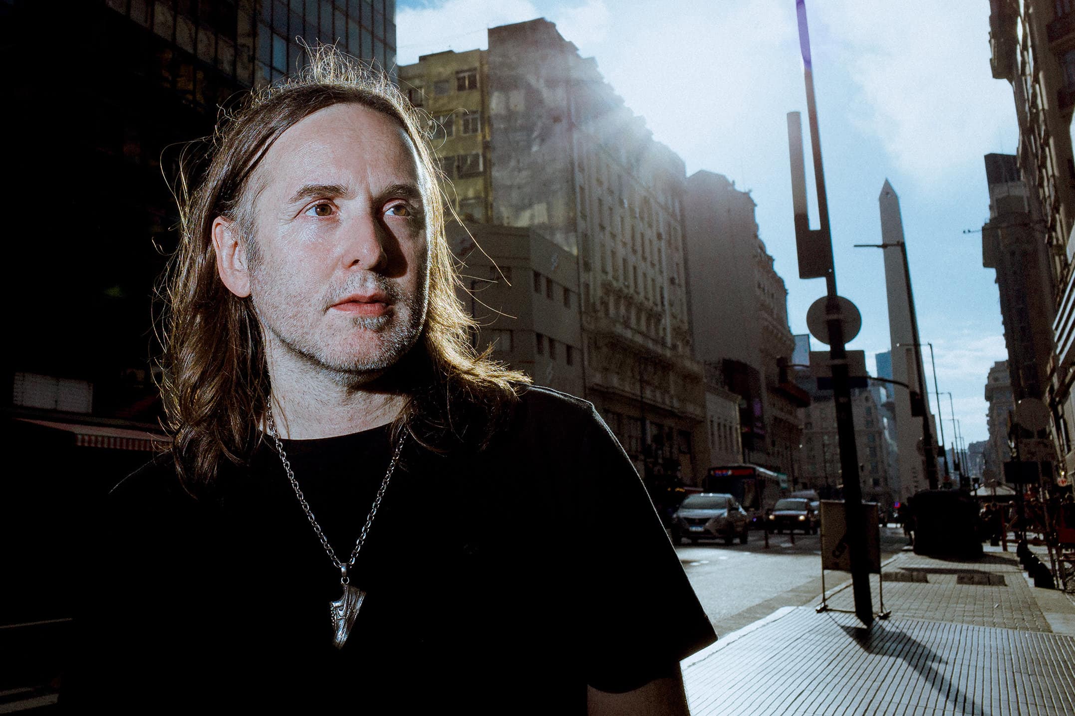 joaquin cubria, a white man in his late 40s, with stubble and shoulder-length brown hair, stands in teh foreground looking to our right. behind him is a busy downtown buenos aires street.