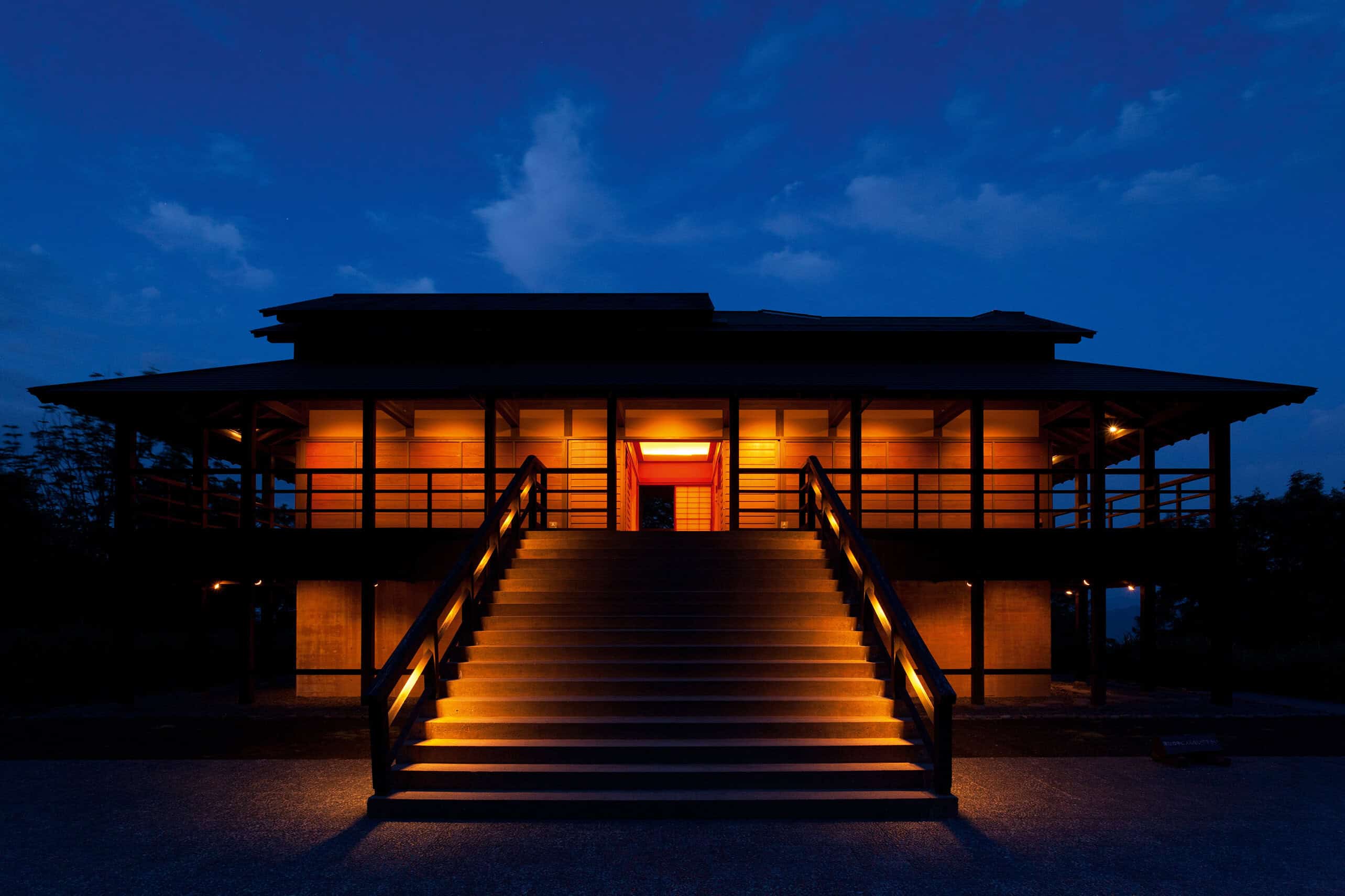 traditional japanese echigo-tsumari region wooden house, lit at night with wam orange light. the house is raised off the ground and has a covered porch area on all sides. a dark, blue night sky is in the background.